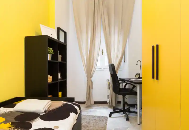 Dormitorio compacto y soleado con cama individual, estantería y un escritorio junto a la ventana; acentos amarillos y cortinas suaves crean un ambiente acogedor.