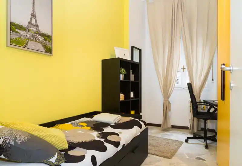 Dormitorio compacto y bien iluminado con cama individual, pared amarilla, estantería y un pequeño escritorio junto a la ventana — espacio acogedor ideal para una persona.