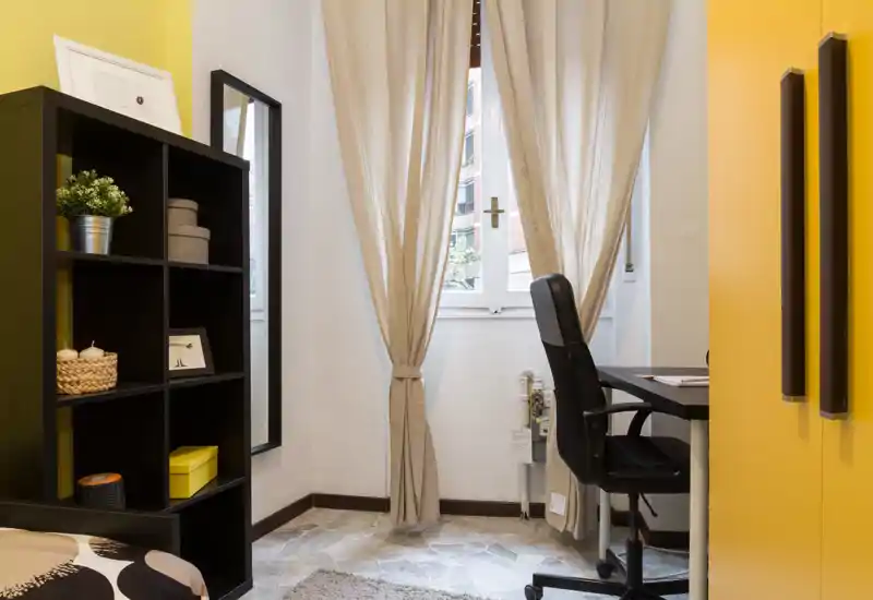 Rincón de dormitorio luminoso y bien decorado con cama parcialmente visible, estantería, escritorio y ventana con cortinas claras — ideal para una persona o zona de estudio.