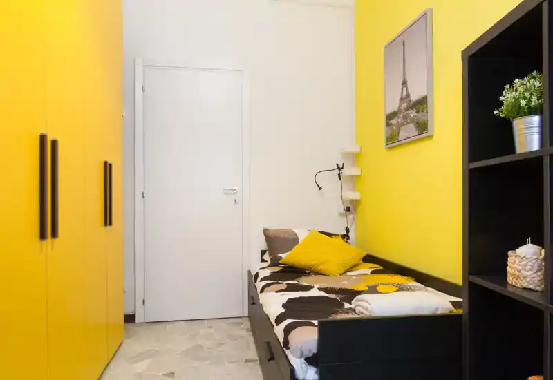 Dormitorio individual luminoso y compacto con estructura de cama negra, ropa de cama estampada y paredes amarillas; un gran armario empotrado y estanterías ofrecen almacenamiento, el ambiente es acogedor y bien decorado.