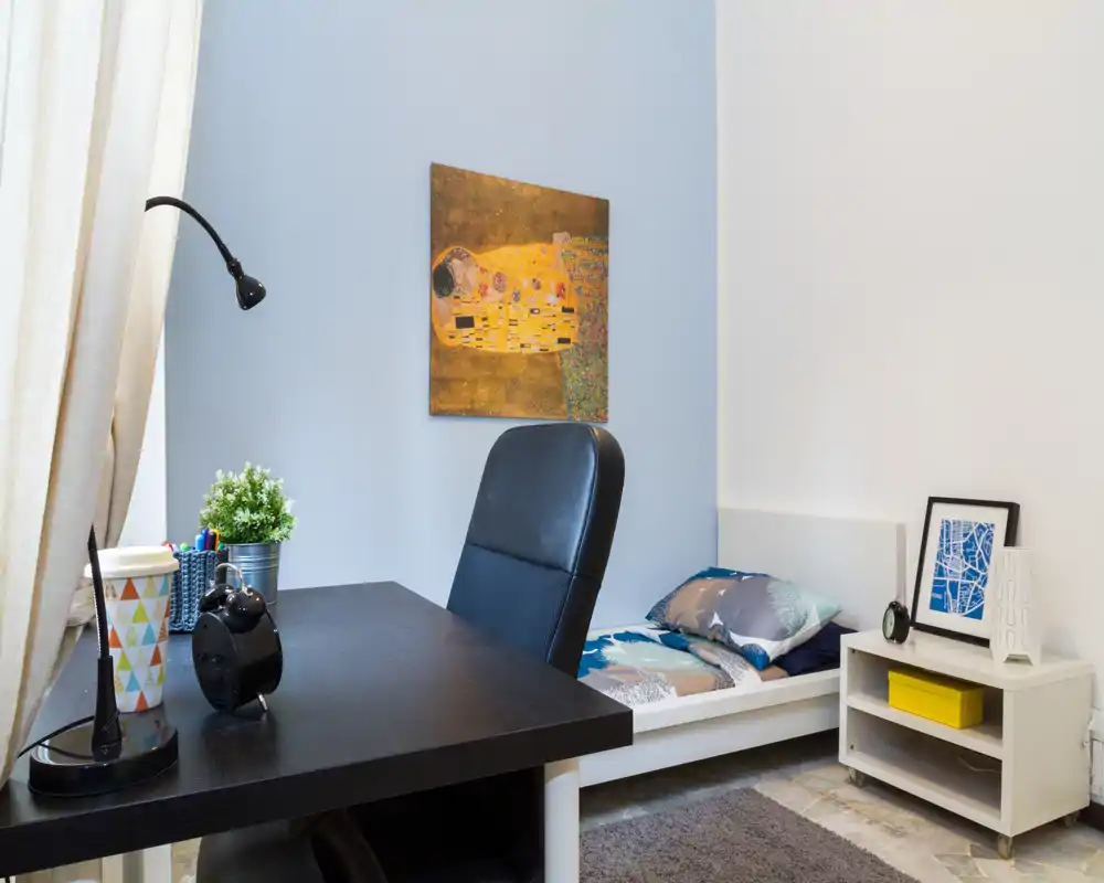 Dormitorio compacto y luminoso con cama individual, mesita y una zona de trabajo en primer plano. Colores neutros, cojines decorativos y cuadro en la pared aportan una sensación ordenada y acogedora, ideal para alquileres de corta estancia.