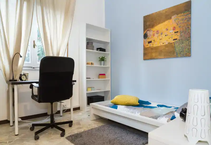 Dormitorio pequeño y bien decorado con cama individual baja, cojín colorido y cuadro; escritorio compacto y silla junto a la ventana que aporta luz natural suave.