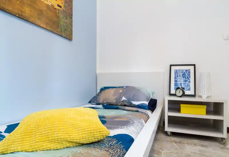 Dormitorio individual moderno y luminoso con ropa de cama estampada, manta amarilla y mesita de noche con lámpara y reloj — ideal para estancias cortas y acogedoras.