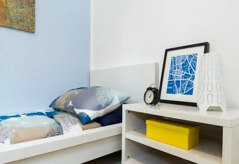 Rincón de dormitorio individual luminoso y ordenado con ropa de cama estampada, mesita con cuadro enmarcado, despertador y lámpara decorativa — un espacio para dormir compacto y bien diseñado.