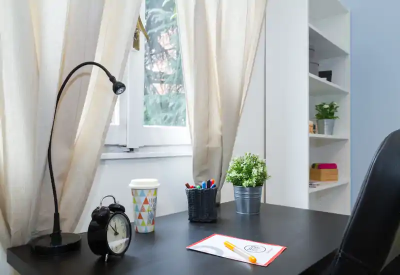 Área de estar/escritorio luminosa y ordenada junto a la ventana con lámpara, reloj, planta en maceta y material de papelería organizado — ideal para trabajar o leer.