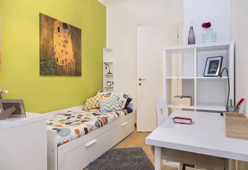 Chambre simple lumineuse et soignée avec lit-coffre blanc, literie colorée et un coin bureau compact — idéale pour étudiant ou célibataire.