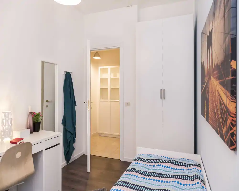 Chambre simple soignée avec lit fait, bureau et armoire ; éclairage chaleureux et style épuré, idéale pour une location confortable.