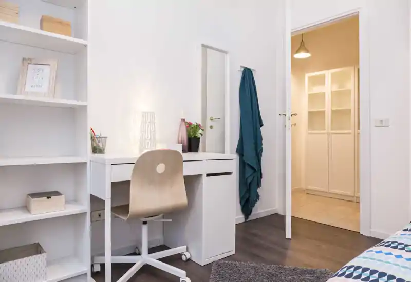 Rincón de dormitorio luminoso y ordenado con escritorio blanco, silla de madera y estantería; se aprecia una pequeña porción de la cama en el borde del encuadre. El espacio es moderno y funcional, con iluminación suave y detalles decorativos cuidados.