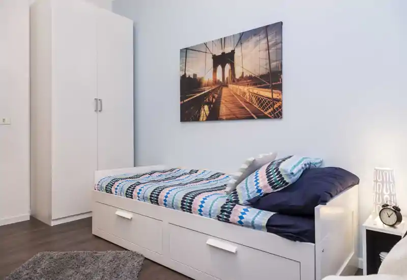 Chambre simple lumineuse et moderne avec un lit-banquette blanc, linge de lit à motifs, armoire et tableau décoratif — parfaite pour une location courte et confortable.