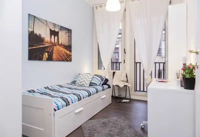Chambre simple lumineuse et soignée avec un lit-banquette blanc, une literie à motifs et une grande fenêtre apportant une belle lumière naturelle — parfaite pour un séjour confortable.