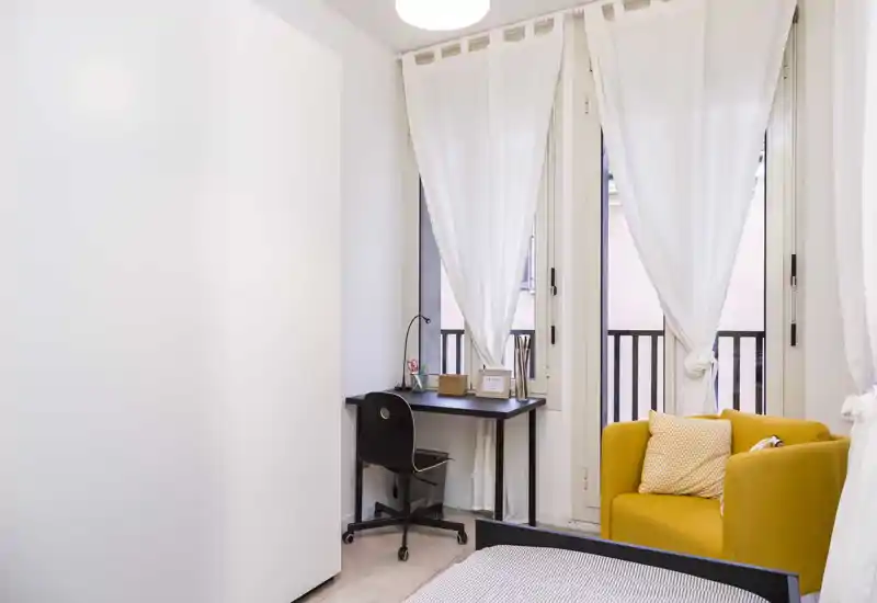 Dormitorio moderno y luminoso con la cama en primer plano, una pequeña zona de trabajo junto a grandes ventanas y un sillón amarillo que aporta calidez.