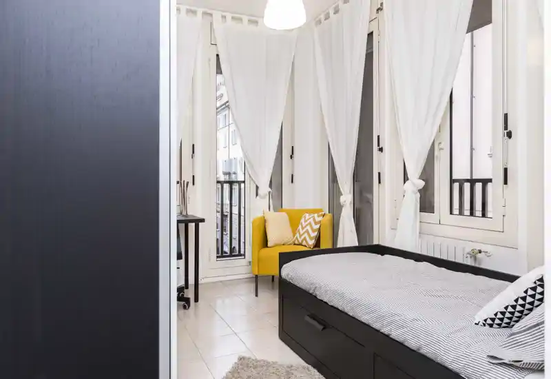 Dormitorio luminoso y moderno con cama individual con cajones, grandes ventanas con cortinas traslúcidas y un acogedor sillón amarillo para crear un rincón de lectura.