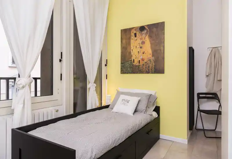 Dormitorio individual luminoso y ordenado con cama tipo daybed de estructura negra, ropa de cama suave y una pared amarilla con cuadro. Grandes ventanas con cortinas blancas proporcionan luz natural y un ambiente acogedor.