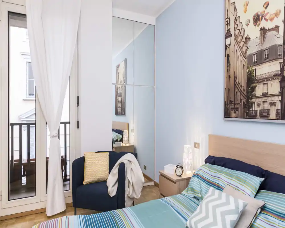 Bright, modern bedroom with a double bed, patterned cushions and a bedside table with lamp; a glass-front wardrobe and a balcony door bring natural light and depth.