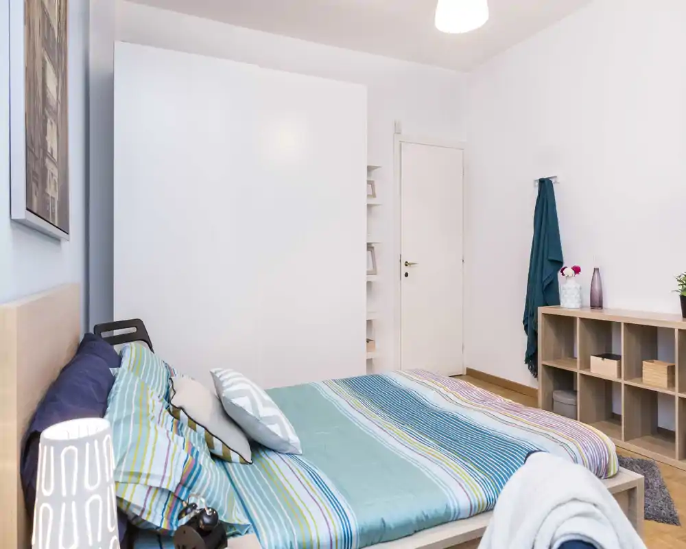 Bright, neatly styled bedroom with a double bed dressed in striped teal linens, a large white wardrobe and open shelving for storage.
