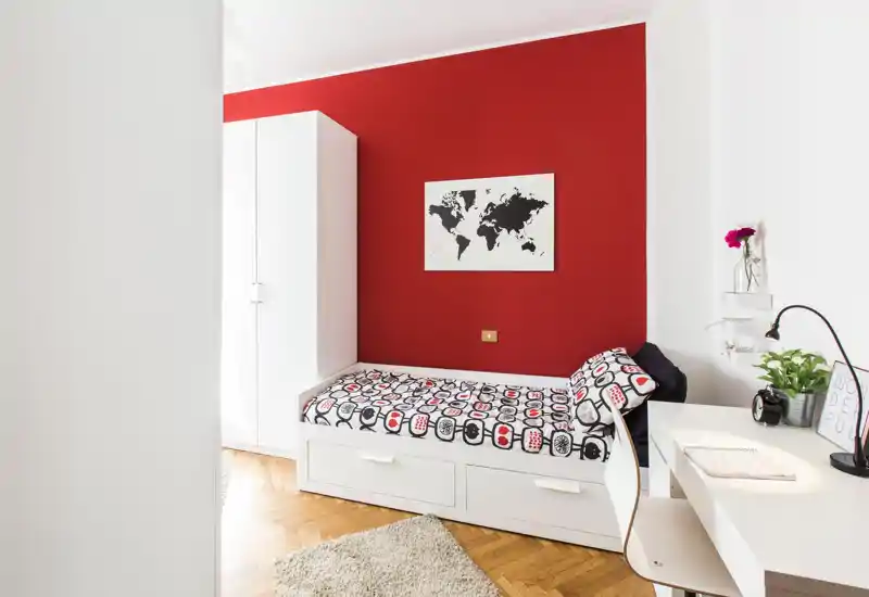 Bright, modern single bedroom with a white daybed, built-in storage drawers and a bold red accent wall. A compact workspace with a desk and plant completes the stylish, functional layout.