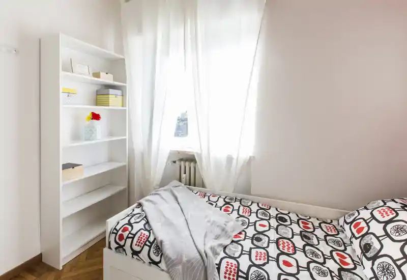 Bright, simply styled single bedroom with patterned bedding, a light throw and a tall white shelving unit beside a window with sheer curtains.