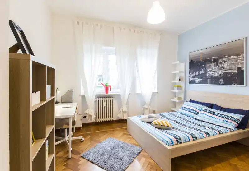 Chambre lumineuse et moderne avec grand lit en bois, linge de lit à motifs et lumière naturelle provenant d'une large fenêtre ; bureau et étagères simples idéaux pour travailler ou se détendre.