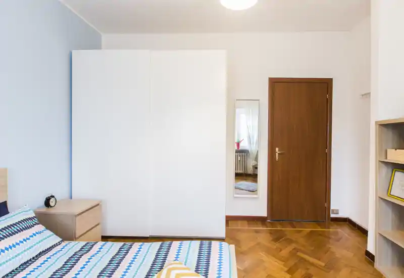 Chambre lumineuse et soignée avec couvre-lit à motifs, table de chevet et grande armoire blanche; parquet propre et lumière chaleureuse.
