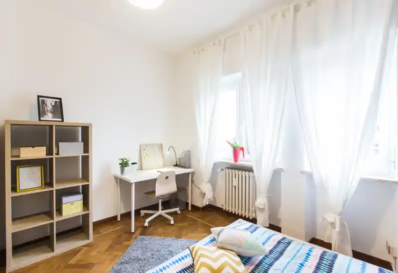 Chambre lumineuse et bien décorée avec lit à motifs, coussins, bureau près de la fenêtre et étagère — idéale pour travailler et se détendre à la lumière naturelle.