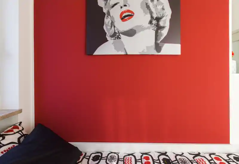Angolo camera da letto compatto con una parete rossa intensa e pop-art moderna sopra il letto; biancheria e cuscini con motivi che danno carattere al piccolo spazio notte.