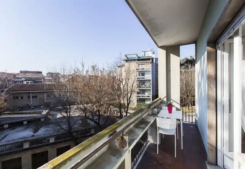 Balcone soleggiato e stretto con un piccolo tavolo e due sedie, con vista su edifici e alberi della città — perfetto per il caffè mattutino.