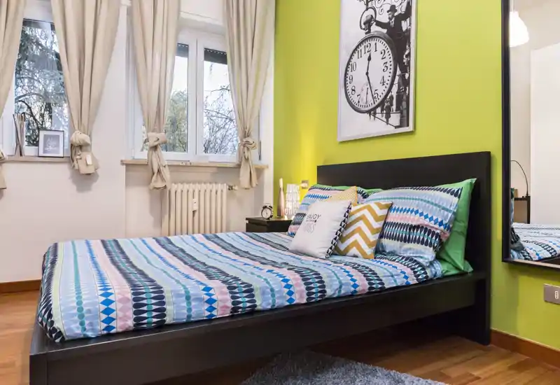 Camera da letto luminosa e moderna con letto matrimoniale colorato, comodino e ampia finestra che introduce luce naturale — perfetta per annunci di affitto.