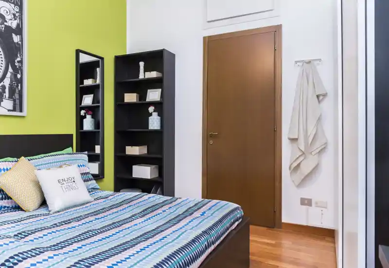 Camera da letto luminosa e curata con un letto matrimoniale a motivo, cuscini decorativi e scaffalature; pavimento in legno caldo e linee pulite creano un'atmosfera accogliente.