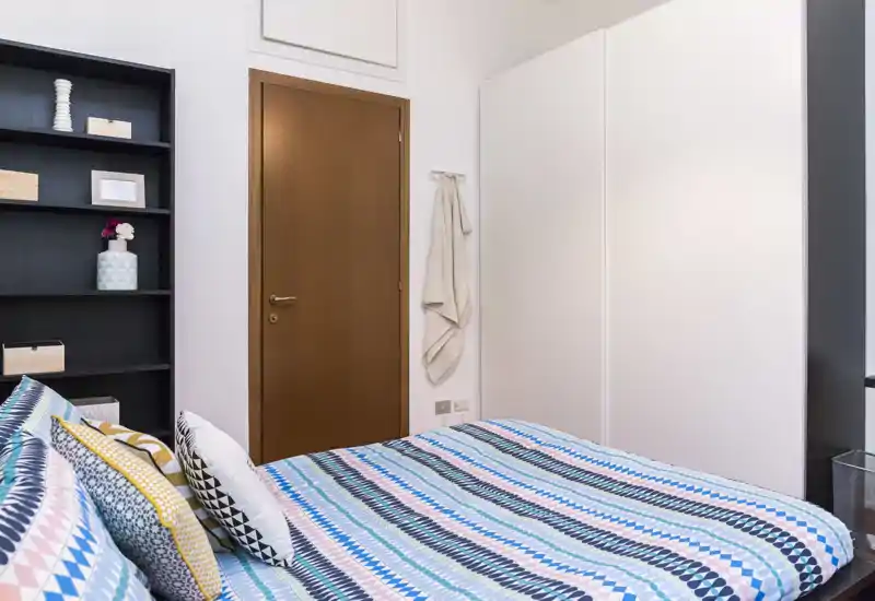 Camera da letto luminosa e ben arredata con un letto matrimoniale decorato in primo piano, un ampio armadio bianco e mensole aperte accanto alla porta. L'ambiente è fresco e ben illuminato con cuscini e tessuti coordinati.