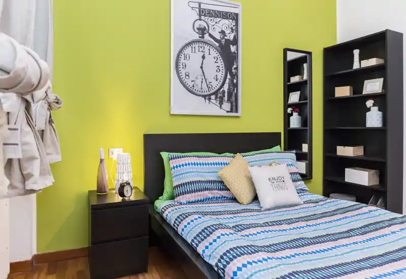 Camera da letto luminosa e curata con letto matrimoniale, biancheria a fantasia e comodino; scaffalatura ordinata e dettagli decorativi rendono l'ambiente accogliente.