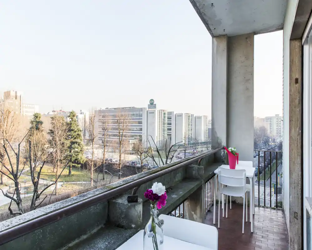 Balcone soleggiato con vista sulla città, piccolo tavolo e sedie e fiori decorativi — ideale per colazioni e momenti di relax all'aperto.
