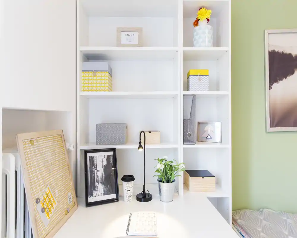 Spazio di lavoro luminoso e ordinato con scaffalature integrate, scatole decorative, lampada da scrivania e una piccola pianta: ideale come angolo studio o home office.