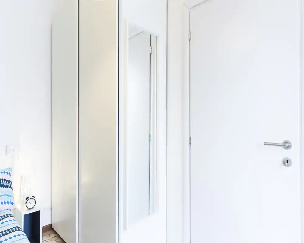 Angolo di camera luminoso e minimal con letto parzialmente visibile, comodino e armadio con specchio a figura intera; pareti bianche e arredi moderni donano una sensazione ariosa.