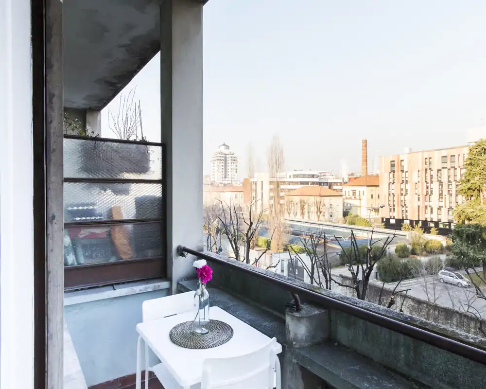 Balcone privato soleggiato con un piccolo tavolo bianco e due sedie, un sottopiatto decorativo e un vaso con fiori, con vista su un tranquillo cortile urbano.