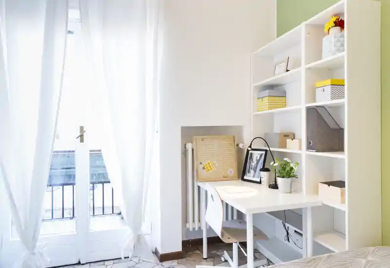 Angolo camera luminoso e ordinato con scrivania bianca e scaffalatura accanto a grandi porte-finestre; tende leggere e scatole decorative creano un'atmosfera ariosa e accogliente.