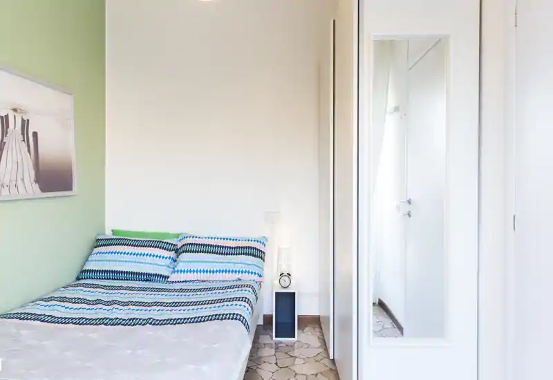 Camera da letto singola luminosa e ordinata con biancheria a fantasia, lampada da comodino e armadio con specchio. L'ambiente è fresco e ben illuminato, perfetto per soggiorni brevi o studenti.