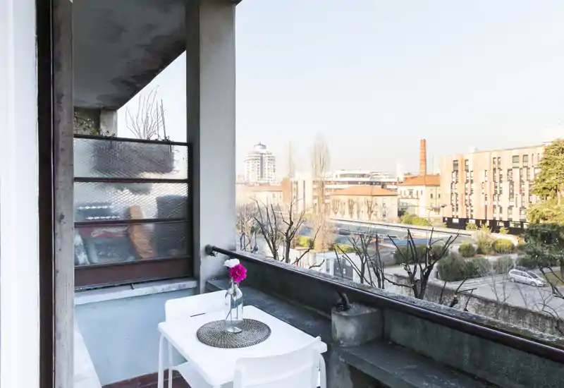 Balcón privado soleado con una pequeña mesa blanca y dos sillas, un salvamanteles decorativo y un jarrón con flores, con vistas a un tranquilo patio urbano.