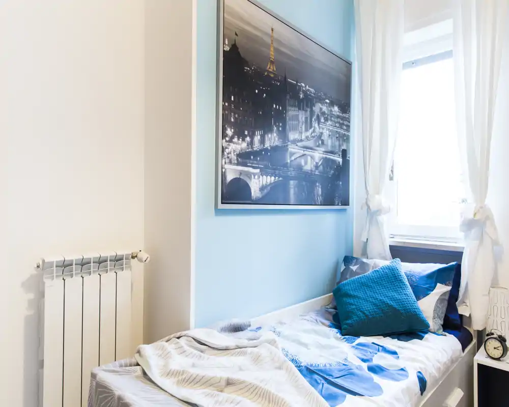 Small, bright single bedroom with a neatly made bed, decorative cushions and a large framed city print above the headboard. Natural light streams through sheer curtains creating a fresh, welcoming atmosphere.