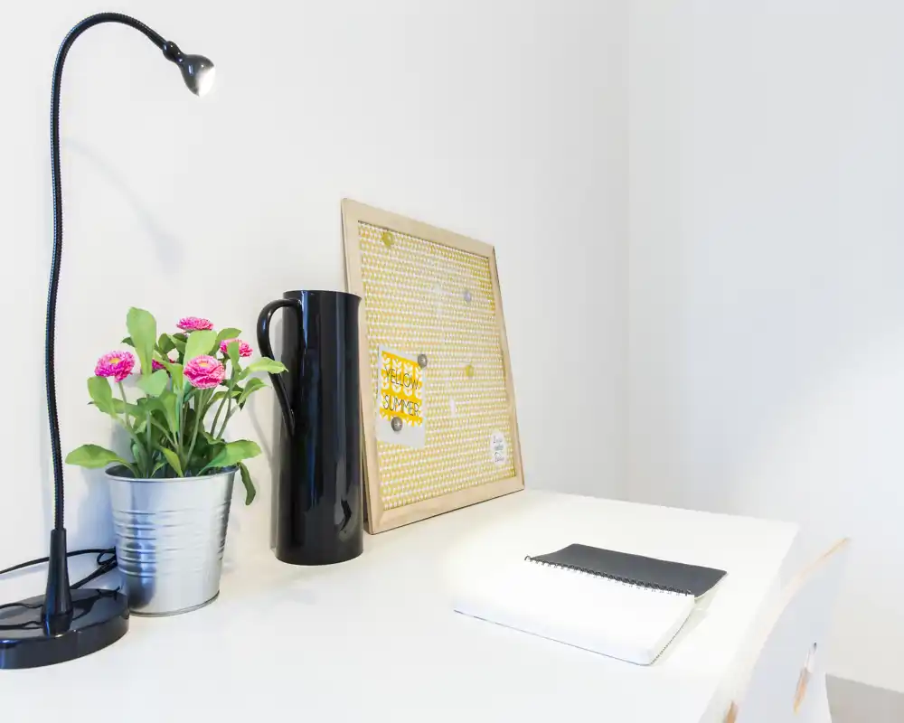 Bright, minimalist desk corner with a lamp, potted plant, decorative board and notebook — a tidy living/working nook ideal for reading or light tasks.