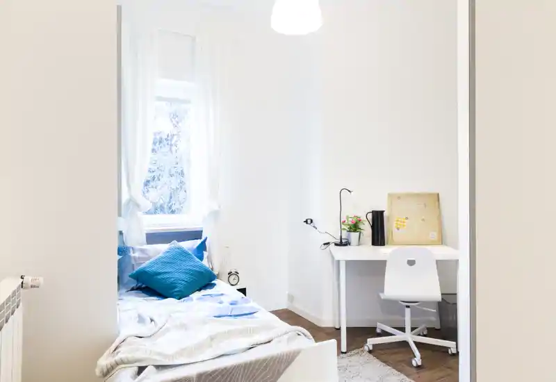 Bright, minimal bedroom with a single bed by the window, blue accent pillows and a small workspace with a white desk and chair.