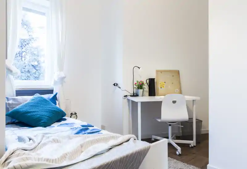 Bright, tidy bedroom featuring a comfortable bed with blue cushions and a compact white desk by the wall—ideal for a student or short-stay rental.