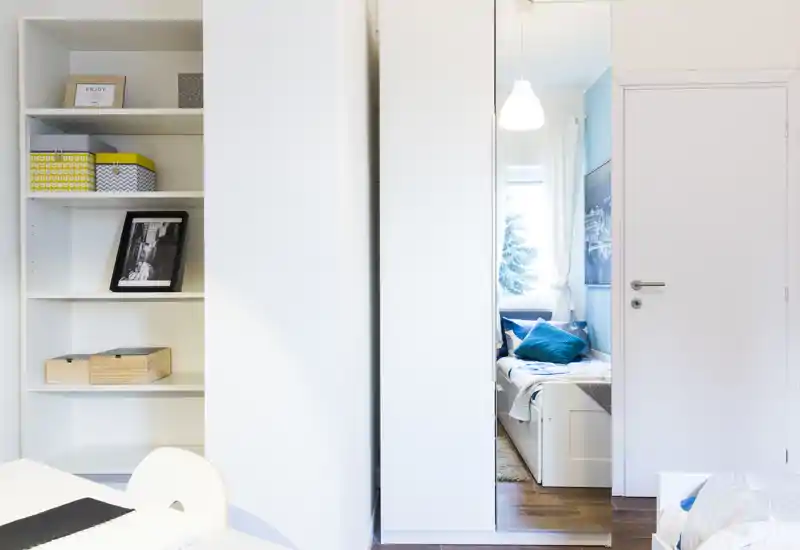 Compact, bright bedroom seen through a doorway and mirrored wardrobe: a neatly made single bed with blue cushions, soft natural light and clean white furnishings.