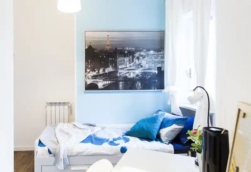 Bright, compact bedroom with a single bed dressed in blue and white linens, decorative cushions and a large framed cityscape above; natural light from the window creates a fresh, inviting atmosphere.