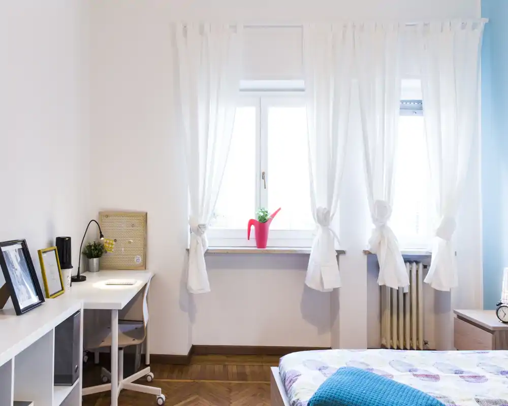 Bright, modern bedroom with a neatly made bed in the foreground, a desk workspace by the window and light, airy curtains letting in natural light.