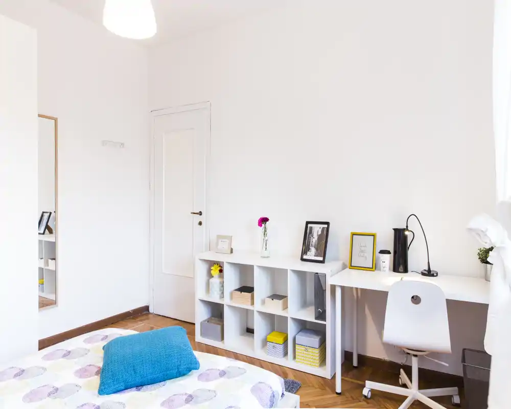 Bright, minimal bedroom with a neatly made bed, a white shelving unit and a small desk — ideal for a tidy, well-lit rental. The room features parquet floors, cheerful accents and a comfortable workspace.