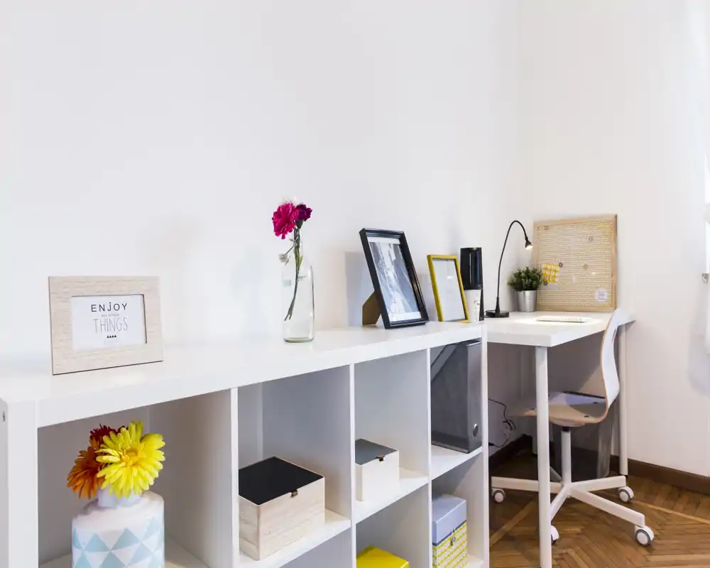 Bright, minimal living/study corner with white shelving, decorative boxes and a compact desk by a window — a neat, functional space ideal for work or reading.