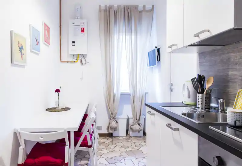 Cocina moderna y compacta con pequeña zona de comedor, muebles blancos, encimera negra y una ventana que aporta luz natural. Detalles decorativos: cuadros, cojines y una flor sobre la mesa.