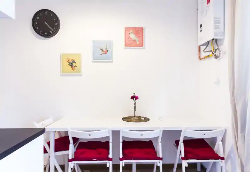 Rincón comedor luminoso y sencillo con mesa blanca, cuatro sillas con cojines rojos y un pequeño centro floral — ideal para comidas en espacios reducidos.