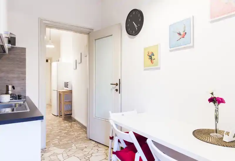 Rincón de cocina compacto y luminoso con fregadero, encimera y una pequeña mesa de comedor. Láminas decorativas, un reloj de pared y flores frescas aportan un ambiente acogedor.