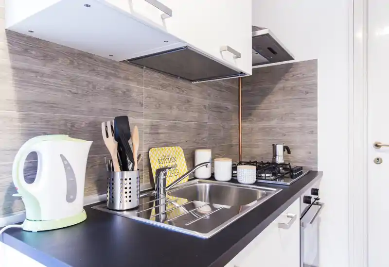 Cocina pequeña y moderna, luminosa, con fregadero de acero inoxidable, cocina de gas y pequeños electrodomésticos; encimera ordenada y salpicadero efecto madera, ideal para alquileres urbanos compactos.